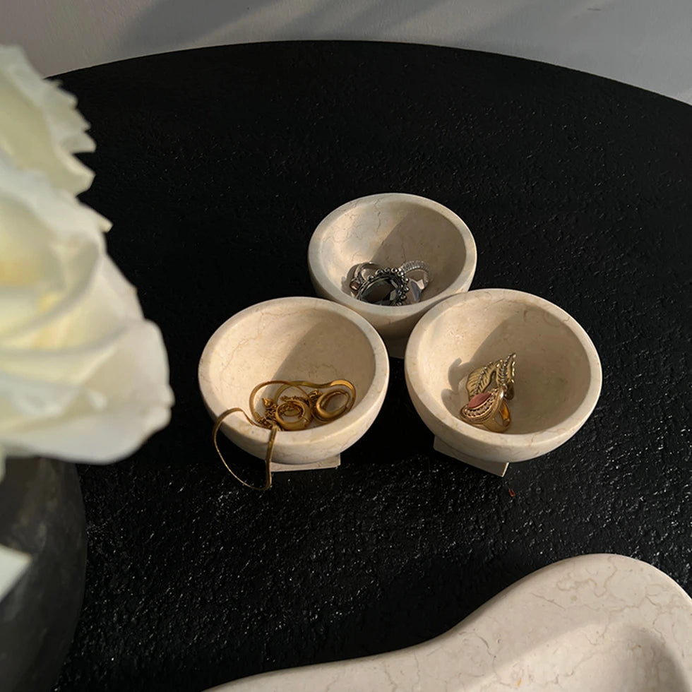 Three small stone bowls on a black table with jewelry inside, next to white flowers.